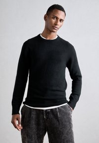 Suéter de punto negro con un escote redondo y puños acanalados, superpuesto a una camisa blanca. Combinado con pantalones de rayas oscuros. Textura suave.