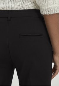 Black tailored trousers feature a smooth fabric, slim fit, and a welt pocket at the back. The waistband is flat with a subtle texture.