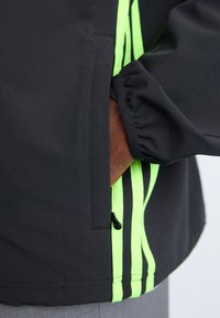 Black jacket with neon green stripes. Features a zippered pocket and elastic cuffs. Smooth texture, contemporary design.