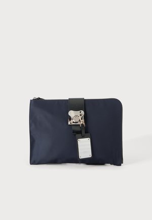 Pochette rectangulaire bleu marine avec bride noire, fermoir métallique argenté et étiquette métallique rectangulaire attachée, sur fond blanc.
