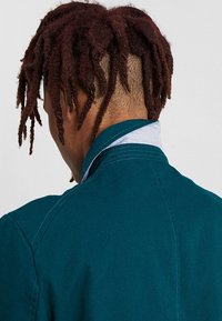 Persona con dreadlocks rojo oscuro y lados rapados lleva una chaqueta azul verdosa y una camisa de cuello azul claro, mirando hacia el lado opuesto de la cámara.