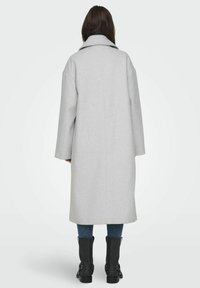 Cappotto di lana grigio chiaro con collo ampio e spalle basse, dotato di maniche lunghe e taglio a lunghezza media, abbinato a stivaletti neri alla caviglia.