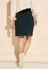 Mörkgrön figursydd kjol med en slät textur, som når till mitten av låret. Kombinerad med en ljus färgad jacka och vita sneakers.