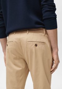 Bež chinos hlače z gladko teksturo, ki imajo dva zadnja žepa z gumbom in tkano pasom. Zgornji del je temno mornarsko modre barve.