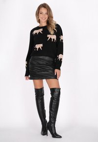 Maglione nero con motivi a tigre beige, abbinato a una minigonna in pelle nera e stivali alti con tacco a spillo neri. Texture lisce in tutto.