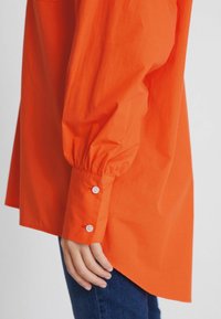 Bras droit dans une chemise à manches longues orange avec des poignets boutonnés, reposant le long du corps vêtu d'un jean bleu.