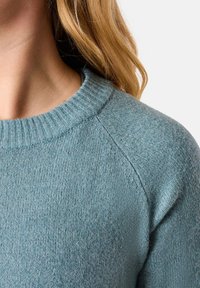 Hellblauer Strickpullover mit einem gerippten runden Halsausschnitt, strukturierter Stoffoberfläche und Raglanärmel. Nahaufnahme zeigt eine glatte Oberflächendetailierung.