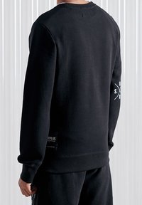 Felpa nera realizzata in materiale spesso con maniche lunghe, con un piccolo logo sulla manica destra e un'etichetta rettangolare sul lato sinistro in basso.