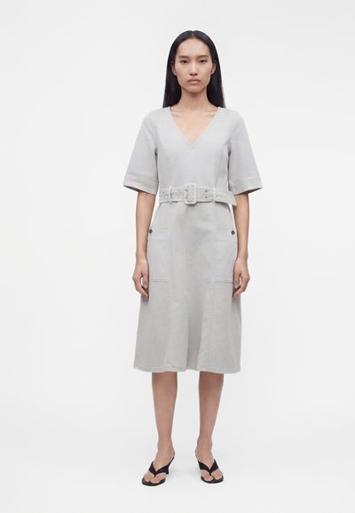 Femme debout portant une robe midi gris clair à col en V, manches aux coudes, taille ceinturée, poches à l'avant et sandales noires à talons.
