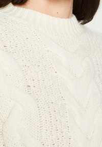 Pull en crème tricoté avec un motif de câble texturé et un col côtelé, présentant une maille lâche et un aspect doux et confortable.