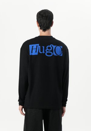 Black long-sleeve shirt with a large blue "Hugo" logo on the back. Cotton material with a relaxed fit and crew neckline.