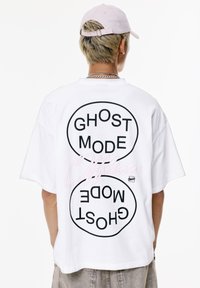 Valkoinen puuvillapaita, jossa on rohkeaa mustaa tekstiä "GHOST MODE" ja "Offline" vaaleanpunaisella, rento malli ja lyhyet hihat.