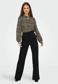 Donna che indossa una blusa a maniche lunghe a strisce zebrate beige e nere con pantaloni neri a vita alta e gamba larga, in piedi con atteggiamento sicuro.