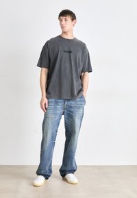 Camiseta de algodón gris de gran tamaño con un logo negro, combinada con jeans de mezclilla azul claro que presentan detalles de patchwork y un corte relajado.