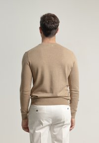 Suéter de punto beig con mangas largas y cuello redondo, combinado con pantalones blancos. La tela del suéter parece suave con una textura sutil.