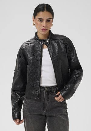 Femme aux cheveux lissés en arrière porte une veste en cuir noire, un débardeur blanc, un jean foncé et des boucles d’oreilles dorées, debout devant un fond uni.
