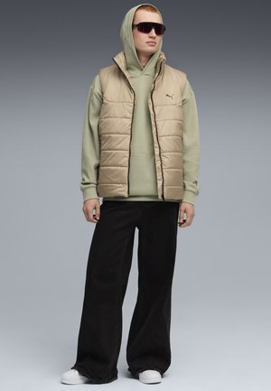 Beige fyllväst över en ljusgrön hoodie, kombinerat med svarta vida jeans och vita sneakers. Modellen bär mörka solglasögon.