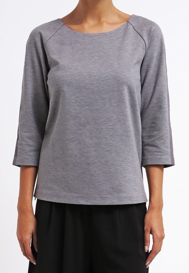 Femme portant un haut gris clair à manches longues trois-quarts et un pantalon noir à jambes larges, debout devant un fond blanc.