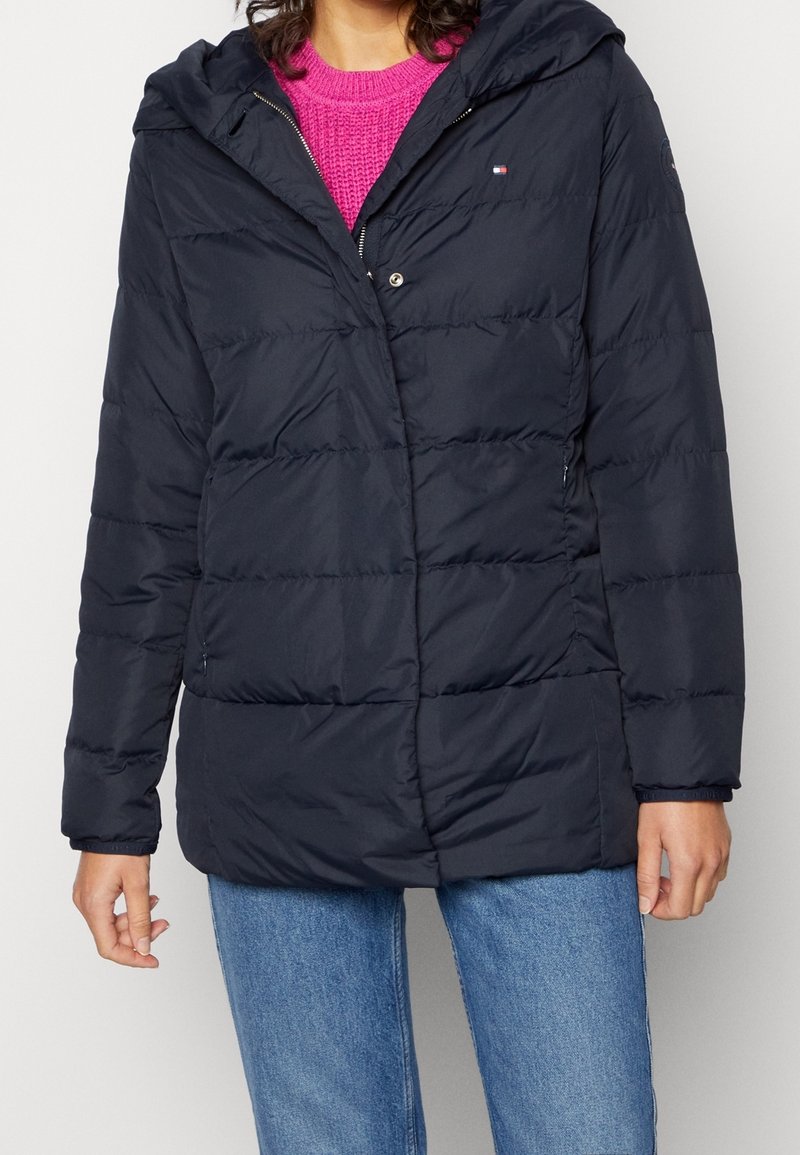 Persona vistiendo una chaqueta acolchada azul marino con capucha sobre un suéter de punto rosa brillante y jeans azules, de pie frente a un fondo liso.