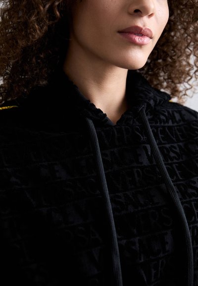Hanorac negru cu un model texturat ce prezintă inscripția "VERSACE". Are glugă, șnur și o textură moale a materialului.