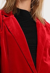 Blazer en velours rouge avec un col cranté, présenté sur un haut noir texturé, mettant en valeur la douce brillance et la coupe ajustée.