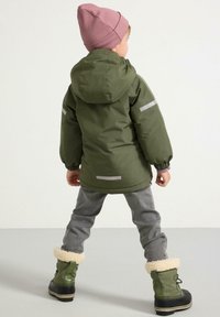 Giacca verde con cappuccio e accenti riflettenti, pantaloni grigi e stivali invernali verdi con bordo in pelliccia, abbinati a un berretto in maglia rosa.