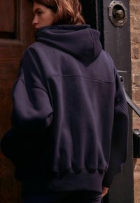 Överdimensionerad marinblå hoodie i mjukt tyg, med en stor huva och ribbad nederkant. Sömdetaljer ger textur. Utsikt bakifrån mot en dörr av trä.