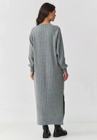 Długą, szaro-rzebrowaną dzianinową sukienkę z długimi rękawami, z bocznymi rozcięciami i okrągłym dekoltem. Miękka tekstura i luźny krój. Widoczne czarne obuwie.