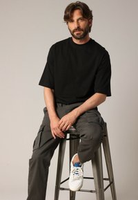 Un homme aux cheveux bruns et à la barbe portant un t-shirt noir, un pantalon cargo gris et des baskets blanches, assis sur un tabouret en métal devant un fond uni.