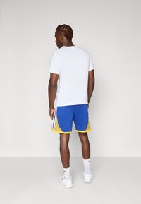 Hvit kortermet t-skjorte i kombinasjon med blå shorts med gule detaljer og stripete sidede detaljer; brukt med hvite sneakers og treningssokker.