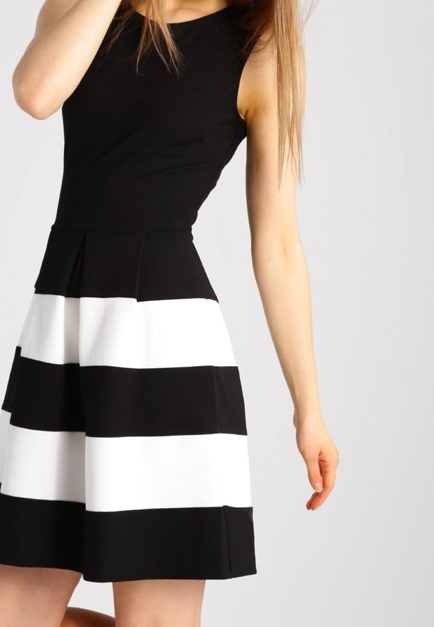 Femme portant une robe noire sans manches avec de larges rayures blanches horizontales sur la jupe, debout devant un fond clair uni.