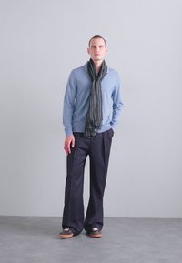 Pulover bleu deschis, eșarfă în dungi întunecate, pantaloni largi de culoare închisă, pantofi maro. Modelul stă pe un fundal gri simplu.