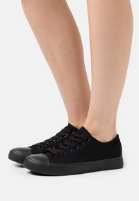 Černé plátěné tenisky s gumovou špičkou, kulatými tkaničkami, nízkým střihem a plochou gumovou podrážkou. Hladká textura s minimálním zdobením.