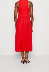 Punainen taitettu mekko, jossa on tiukka ribbausvarsi ja polvipituus. Materiaali näyttää kevyeltä. Siinä on takana vetskari ja minimaalinen yksityiskohta.