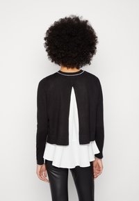 Cárdigan negro con un diseño de espalda dividida, textura de canalé y costuras grises en contraste en el cuello y el dobladillo, llevado sobre una blusa peplum blanca.