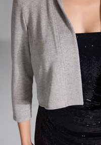 Cardigan grigio accorciato con un leggero scintillio, caratterizzato da una superficie liscia e un design a apertura frontale. Indossato sopra un abito nero con paillettes.