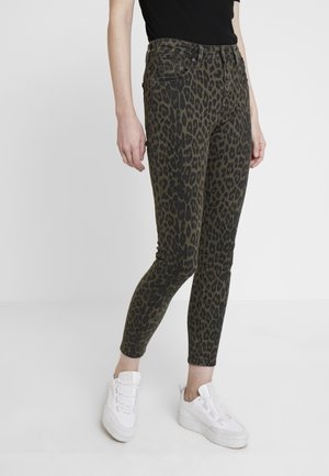 Persoon draagt olijfgroene skinny jeans met zwarte luipaardprint en witte plateau sneakers, staand tegen een effen witte achtergrond.