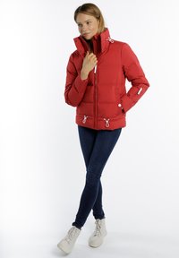 Rode pufferjack met een hoge kraag, ritssluiting aan de voorkant en een trekkoord onderaan. Heeft horizontale stiksels en zijvakjes. Gekozen in combinatie met donkere jeans.