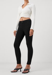 Jeans negros ajustados combinados con un top corto de canalé blanco. Con cierre de botón y mangas largas. La modelo lleva sandalias de tacón negras.