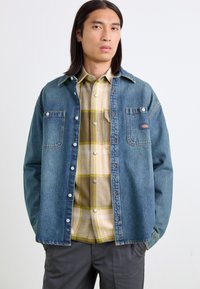 Denimskjorte i blå, med to brystlommer og metalknapper. Bæres over en lys ternet skjorte med gule og beige nuancer.