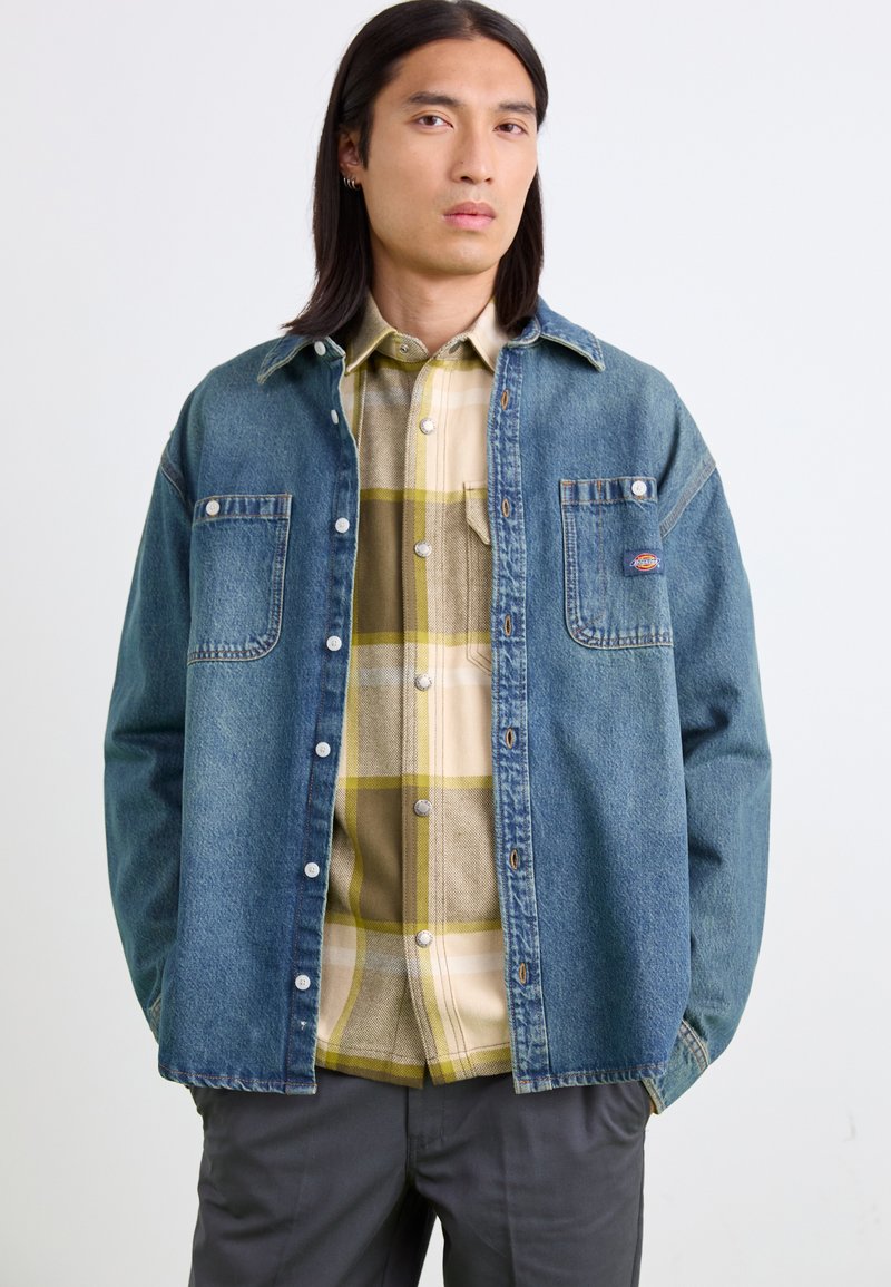 Denimskjorte i blå, med to brystlommer og metalknapper. Bæres over en lys ternet skjorte med gule og beige nuancer.