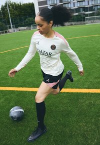 A sportos öltözék egy fehér hosszúujjú mezből áll, rózsaszín részletekkel és logóval, mely fekete rövidnadrággal és térd fölé érő zoknival van párosítva. Egy fekete focilabda található a pályán.
