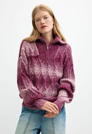 Junge Frau mit langen blonden Haaren, die einen lila und pink gemusterten Strickpullover und hellblaue Jeans vor weißem Hintergrund trägt.