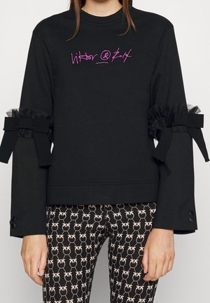 Zwarte top met lange mouwen met een roze logo in sierletters, strik- en ruchesdetails op de mouwen, gecombineerd met zwarte broek met patroon en witte bloemenprint.