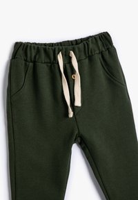 Pantalones de cintura elástica en verde oscuro, con cordones de algodón en color crema, bolsillos laterales, y un pequeño detalle de botón marrón en la parte frontal.