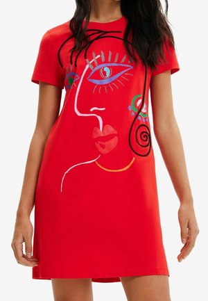 Mujer que lleva un vestido rojo de manga corta con un diseño abstracto y colorido de un rostro en la parte delantera, con ojos, labios y remolinos.