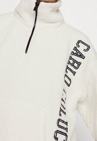 Pullover in pile bianco con colletto alto e zip a metà. Presenta testo ricamato nero lungo le maniche e una tasca frontale. Texture morbida.