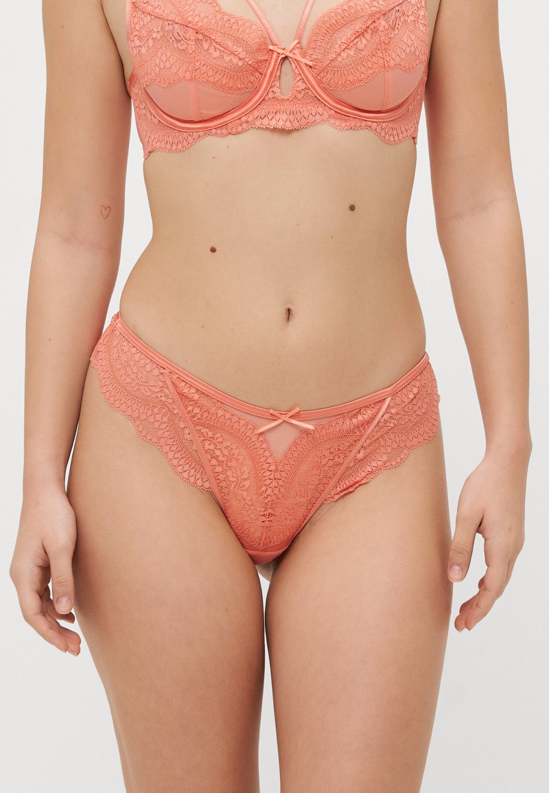 Korallivärinen pitsialusvaatesetti, johon kuuluu bralette, jossa on monimutkaiset aallotetut reunat, sekä yhteensopivat alushousut, joissa on hieno nauha edessä.
