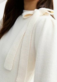 Biały sweter o teksturowanym wykończeniu, z wysokim kołnierzem i miękką tkaniną. Charakterystyczne detale to długie pasy materiału przy dekolcie.