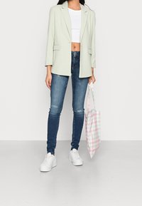 Chaqueta corta de un verde claro, camiseta blanca, jeans skinny de un azul oscuro, zapatillas blancas y una bolsa tipo tote a rayas en rosa y verde.